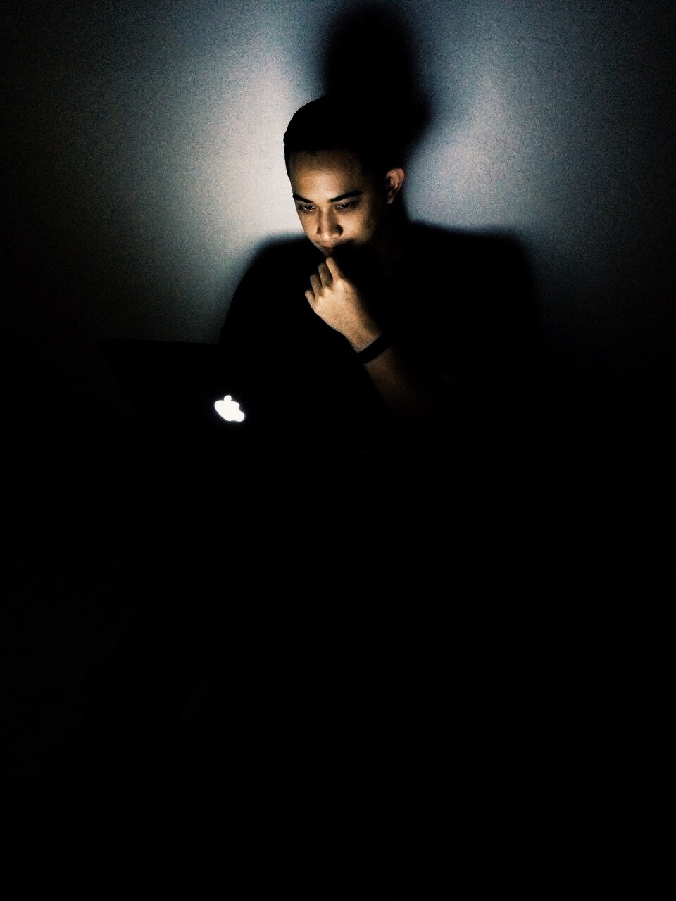 A person illuminated only by a glowing MacBook screen in a dark room, chin resting on hand in a thoughtful pose.